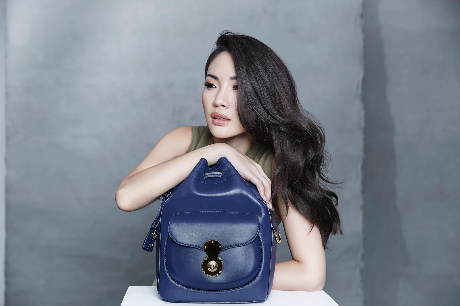 Woman with long dark hair leaning on a navy blue leather bucket bag in a studio setting