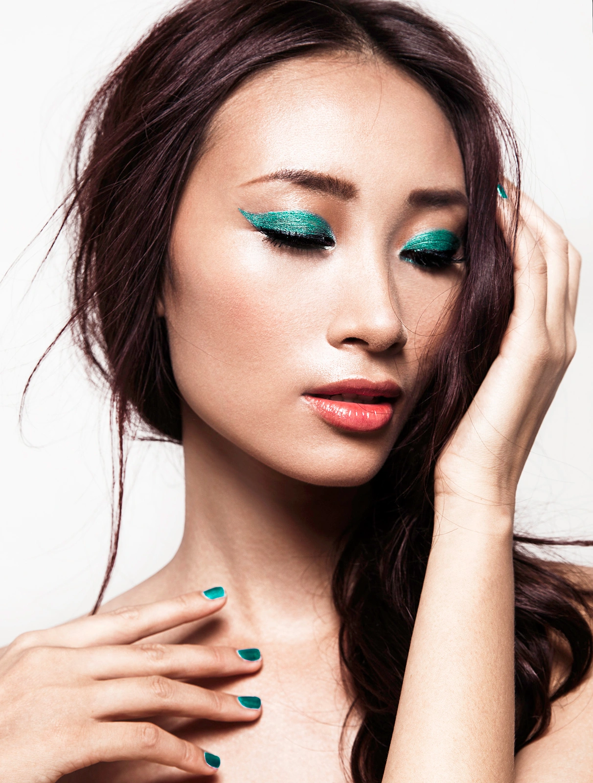 Close-up portrait of a woman with vibrant teal eye makeup, glossy dark hair, and matching teal nail polish against a plain white background.