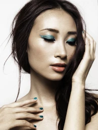 Close-up portrait of a woman with vibrant teal eye makeup, glossy dark hair, and matching teal nail polish against a plain white background.