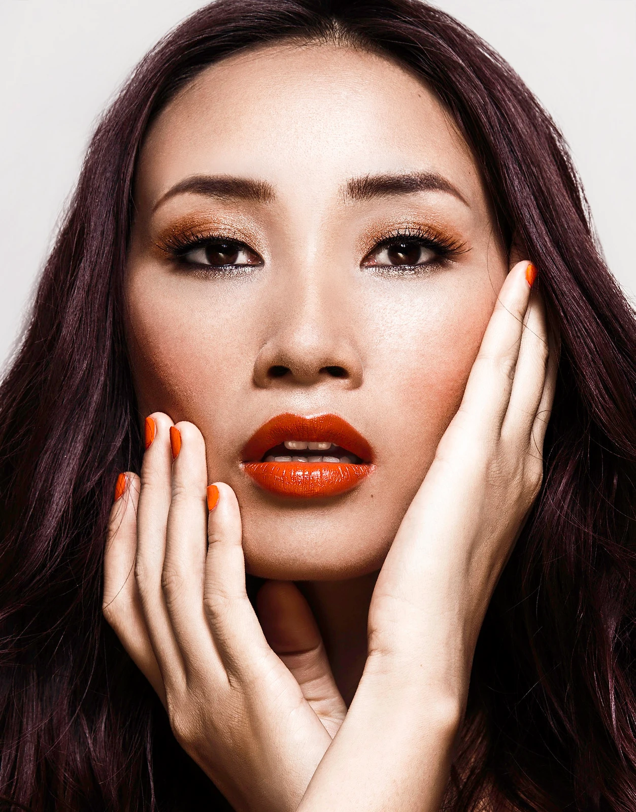 Close-up beauty portrait of an East Asian woman with dark hair, orange lipstick, and vibrant orange nails framing her face.
