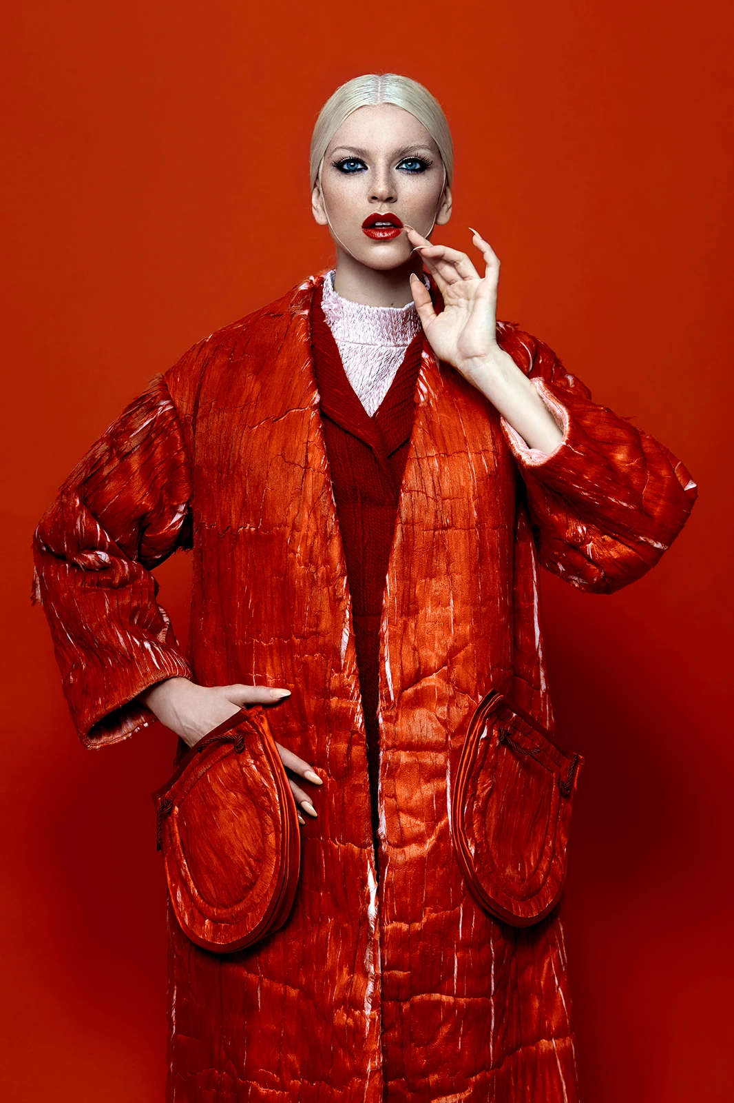 Model with slicked blonde hair wearing a textured red coat against a matching red seamless studio backdrop.