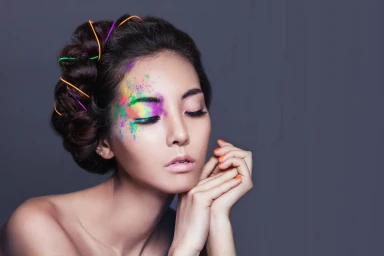 Woman with braided dark hair woven with colourful wires, vibrant multi-coloured powder makeup around one eye, and hands posed near face.