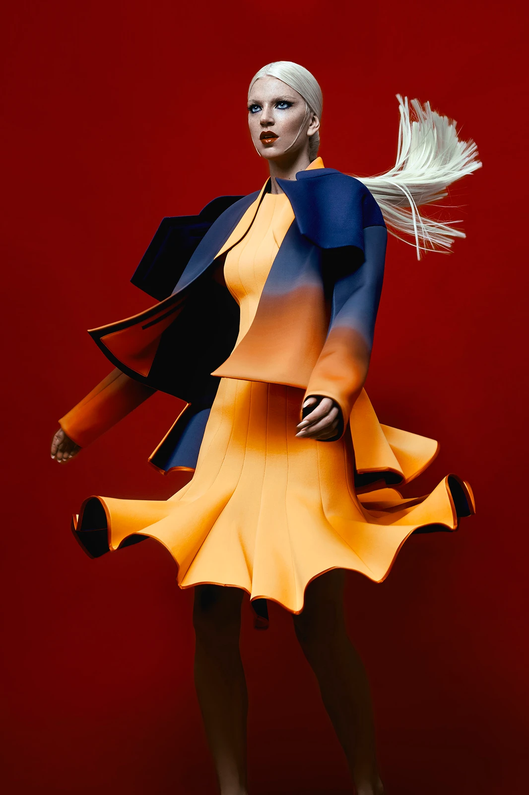 Model with platinum blonde hair in motion wearing an orange dress and blue jacket against a red background