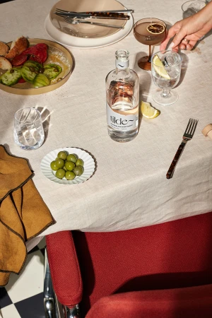 Overhead view of a dining table with a glass bottle of Tilde spirits, appetizers, glasses, and a red armchair in the foreground.