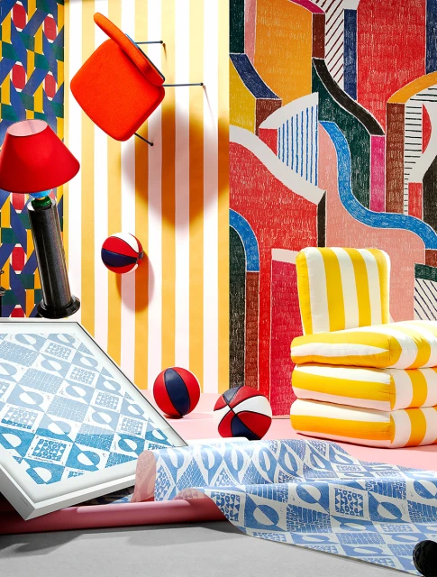 Still life composition with a floating orange chair, yellow striped cushions, balls, and patterned fabrics against colorful backgrounds.