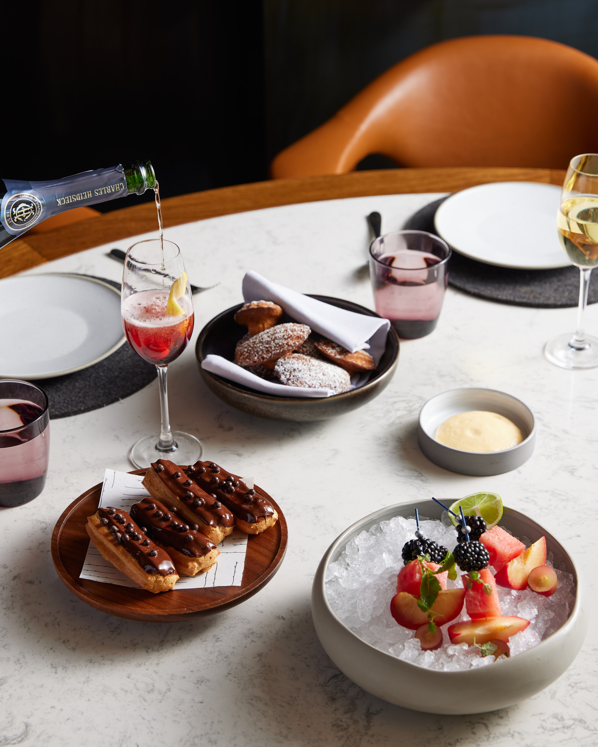 Elegant table setting with champagne pouring into a flute, surrounded by pastries, fruit, and drinks, evoking a refined culinary experience.