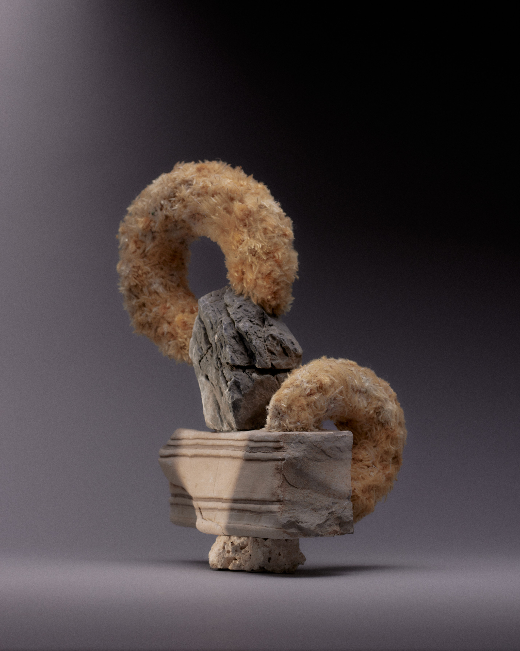 Abstract still life arrangement featuring donuts and rocks on a pedestal, in a studio with soft lighting and a dark gray background.