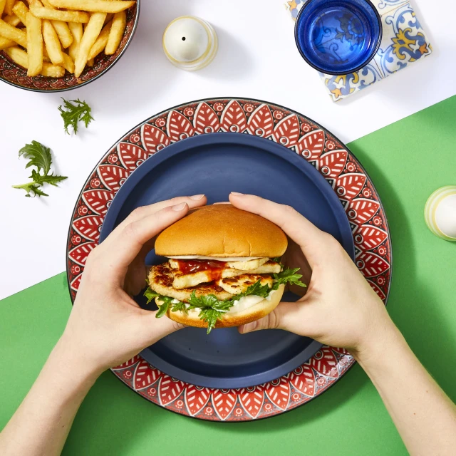 Overhead shot of a burger on a plate held by two hands, set against a colorful background with fries and a drink nearby.