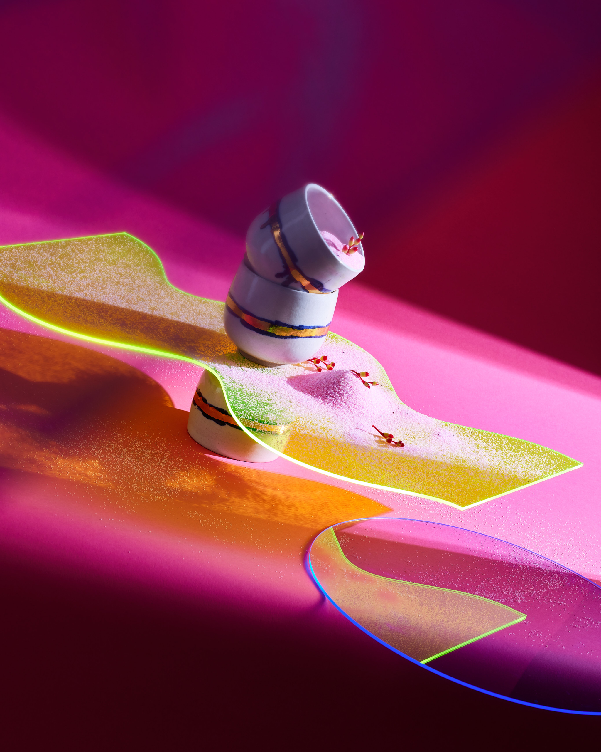Still life of stacked white bowls with gold trim on a yellow platform with pink powder against a vibrant pink background and soft studio lighting.