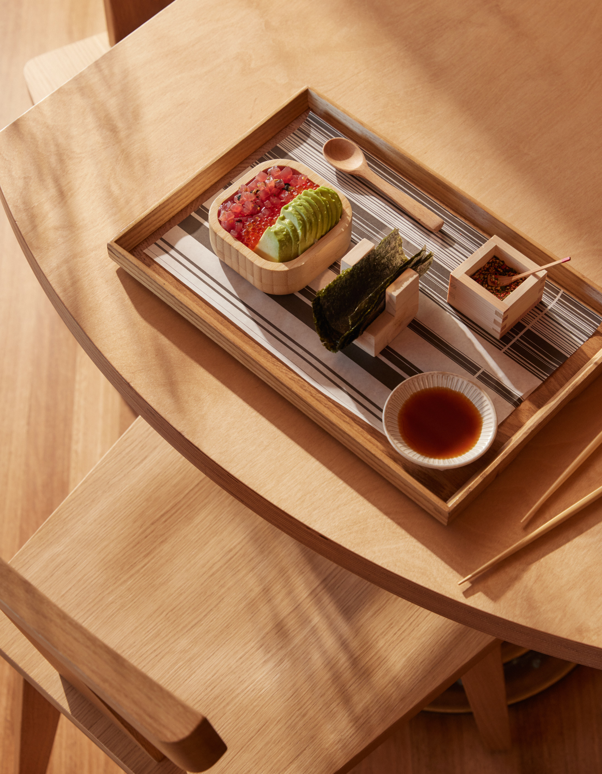 Overhead shot of sushi ingredients arranged on a wooden tray with avocado, tuna, seaweed, soy sauce, and chopsticks on a wooden table.