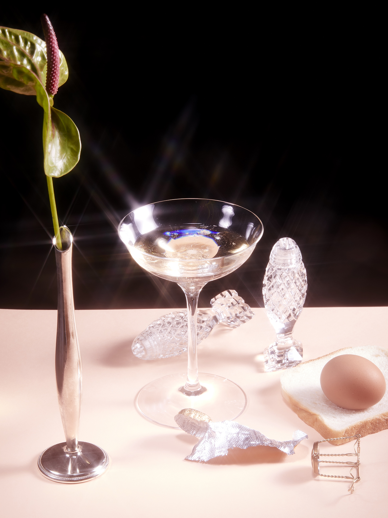 Still life: champagne coupe with anthurium, egg on toast, and crystal salt shakers on a peach-colored table against a black backdrop.