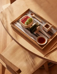 Overhead food photography of a wooden tray with a small bowl of poke, sliced avocado, nori sheets, and a soy sauce dish on a wooden table.