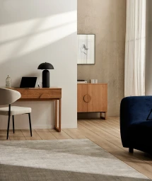 Modern minimalist home office setup with a wooden desk, black lamp, laptop, and a cream chair in a sunlit room.