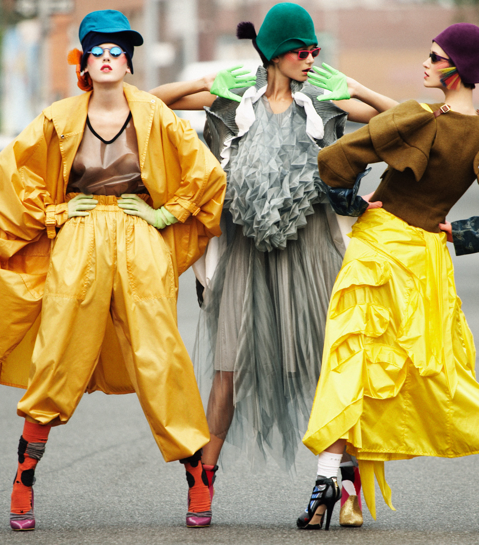 Three women in colorful, avant-garde high fashion stand on an asphalt road, posing in brightly colored outfits, hats, and gloves.