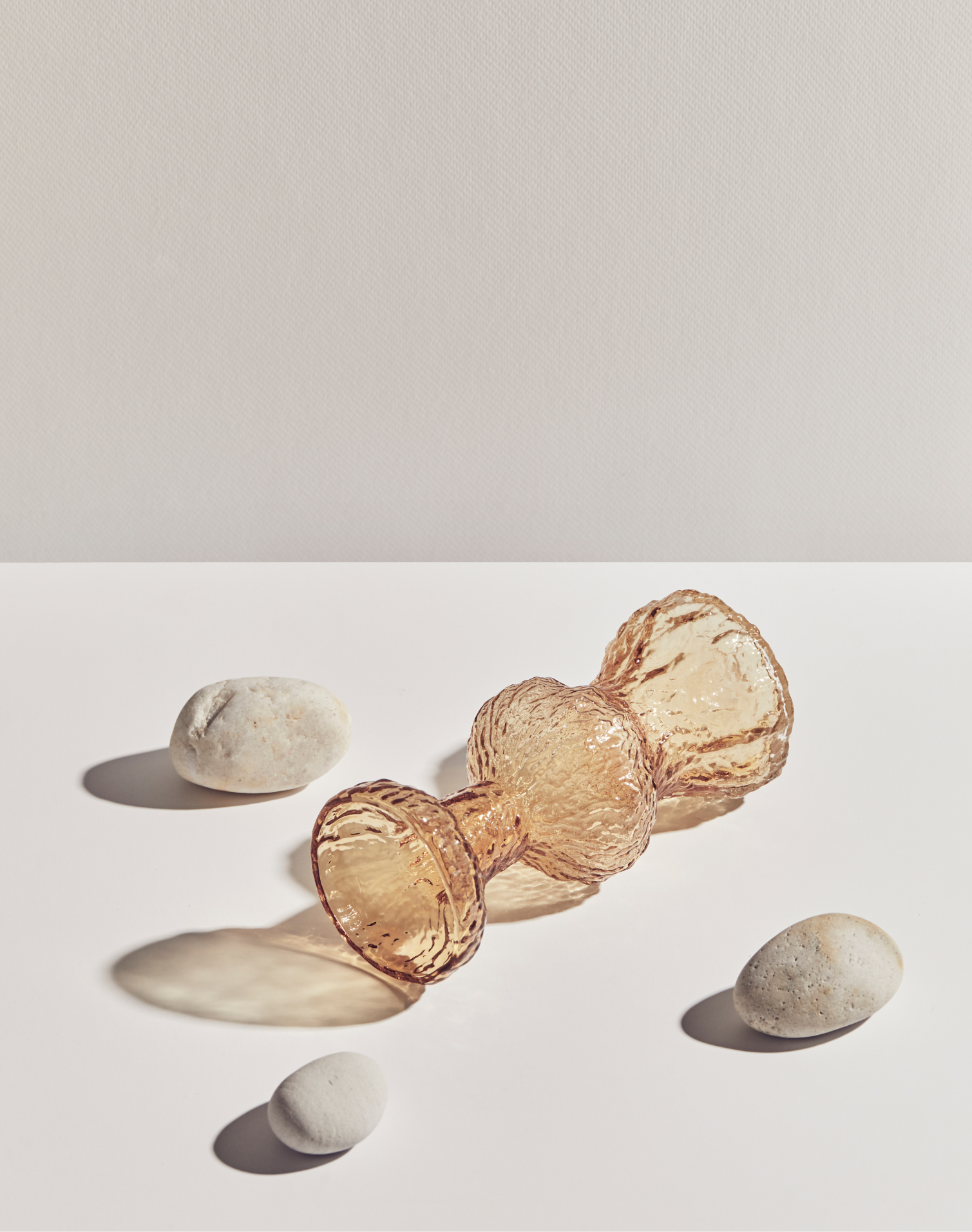 Artistic product shot of an amber glass vase lying on its side, surrounded by three round rocks on a white background with a cream backdrop.