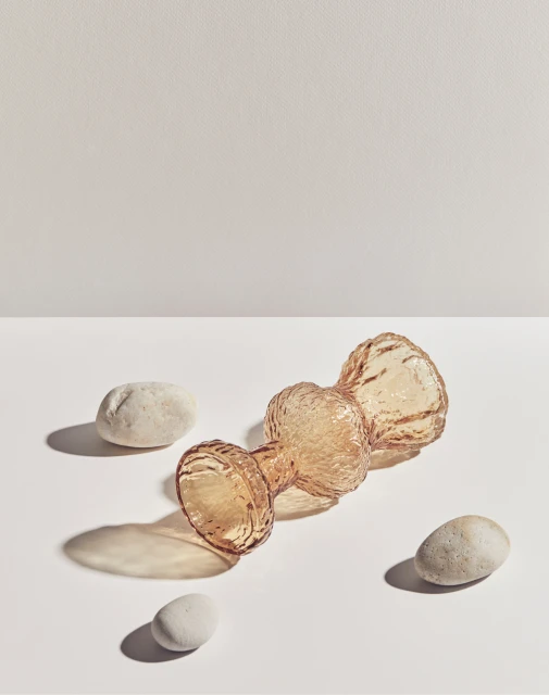 Artistic product shot of an amber glass vase lying on its side, surrounded by three round rocks on a white background with a cream backdrop.