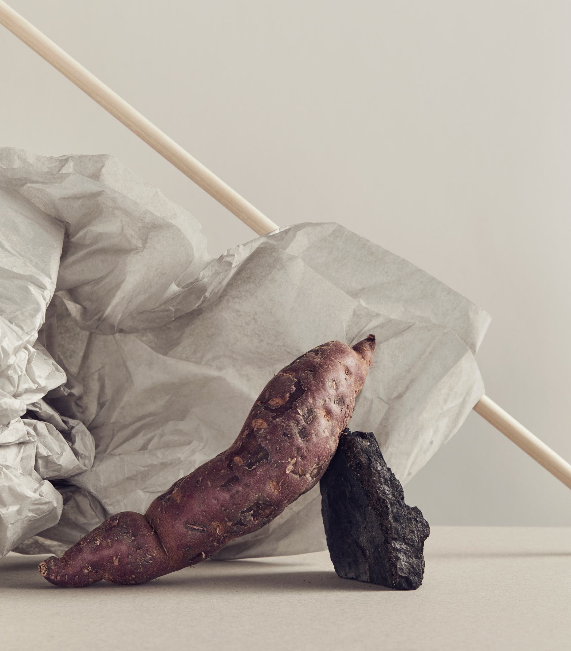 Still life photograph of a yam with textured white paper and a piece of black charcoal on a beige surface in a studio setting.