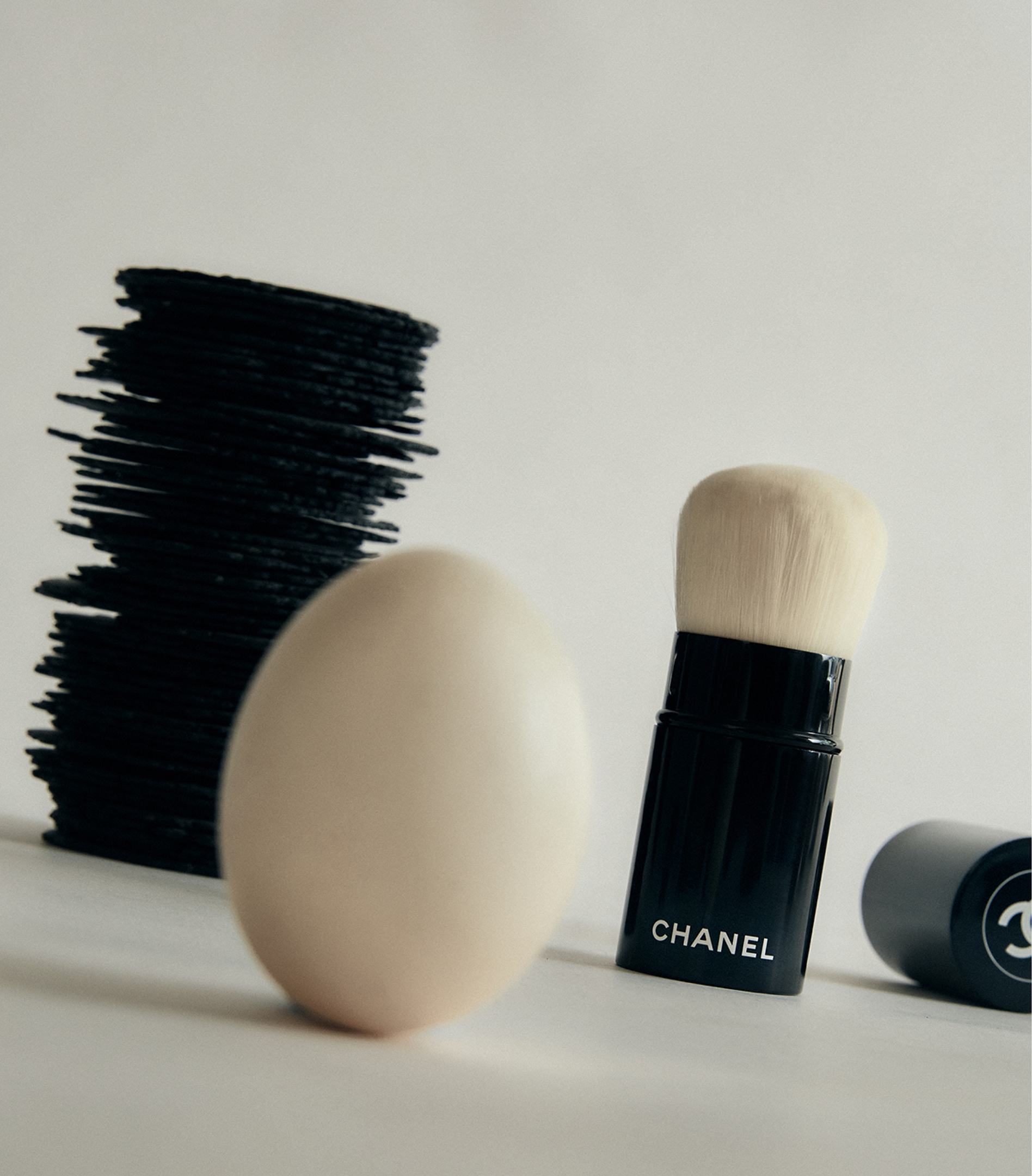 Still life of black makeup applicators, a white chicken egg, and a black Chanel makeup brush on a white surface.