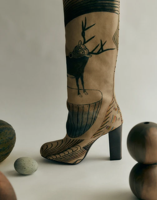 High-heeled boot with deer art, presented against a neutral background, accompanied by round stones. Fashion editorial concept.