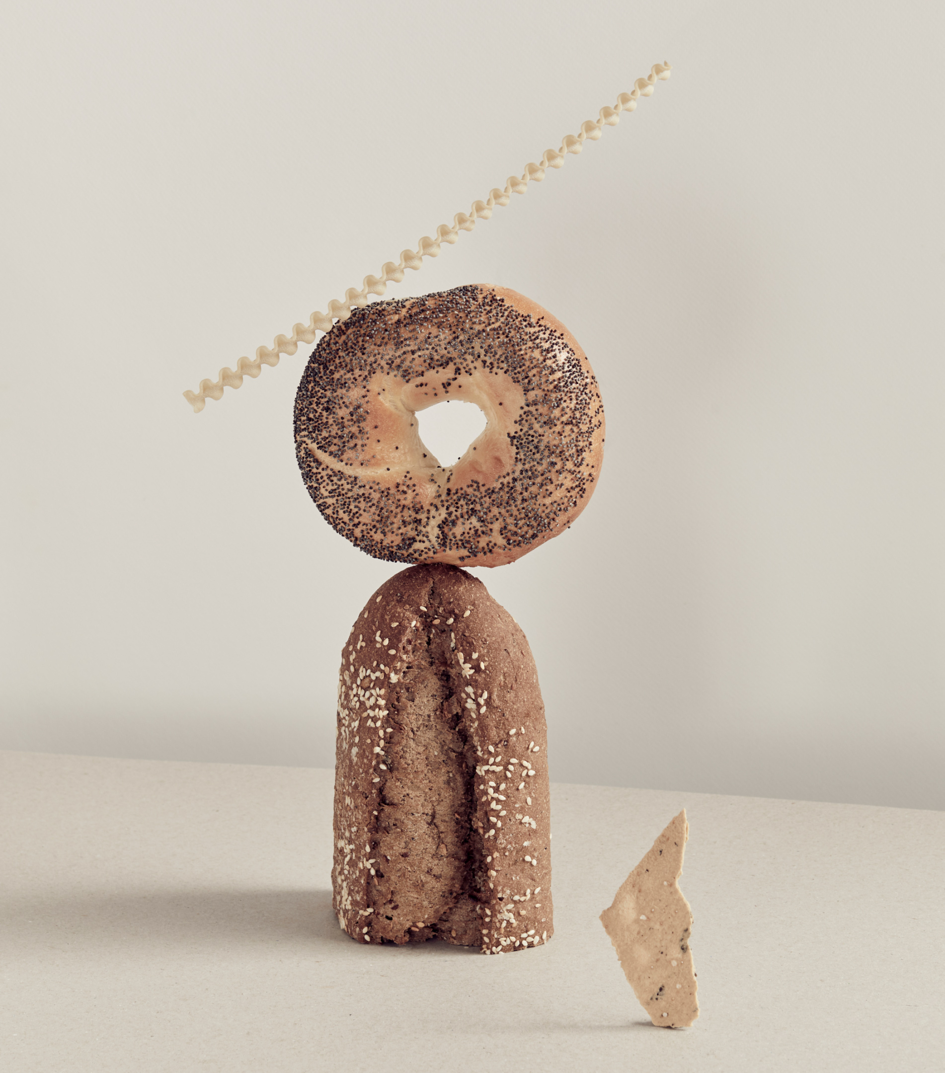 Still life of stacked baked goods: a bagel, a loaf of bread, pasta strands, and a piece of baked good set against a neutral background.