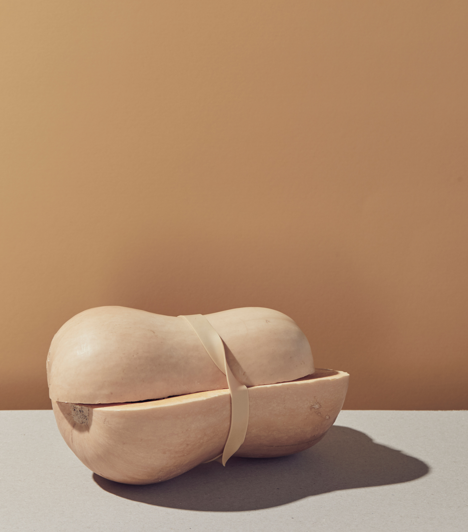 Still life of a halved butternut squash tied with ribbon on a grey surface against a tan background in bright studio lighting.