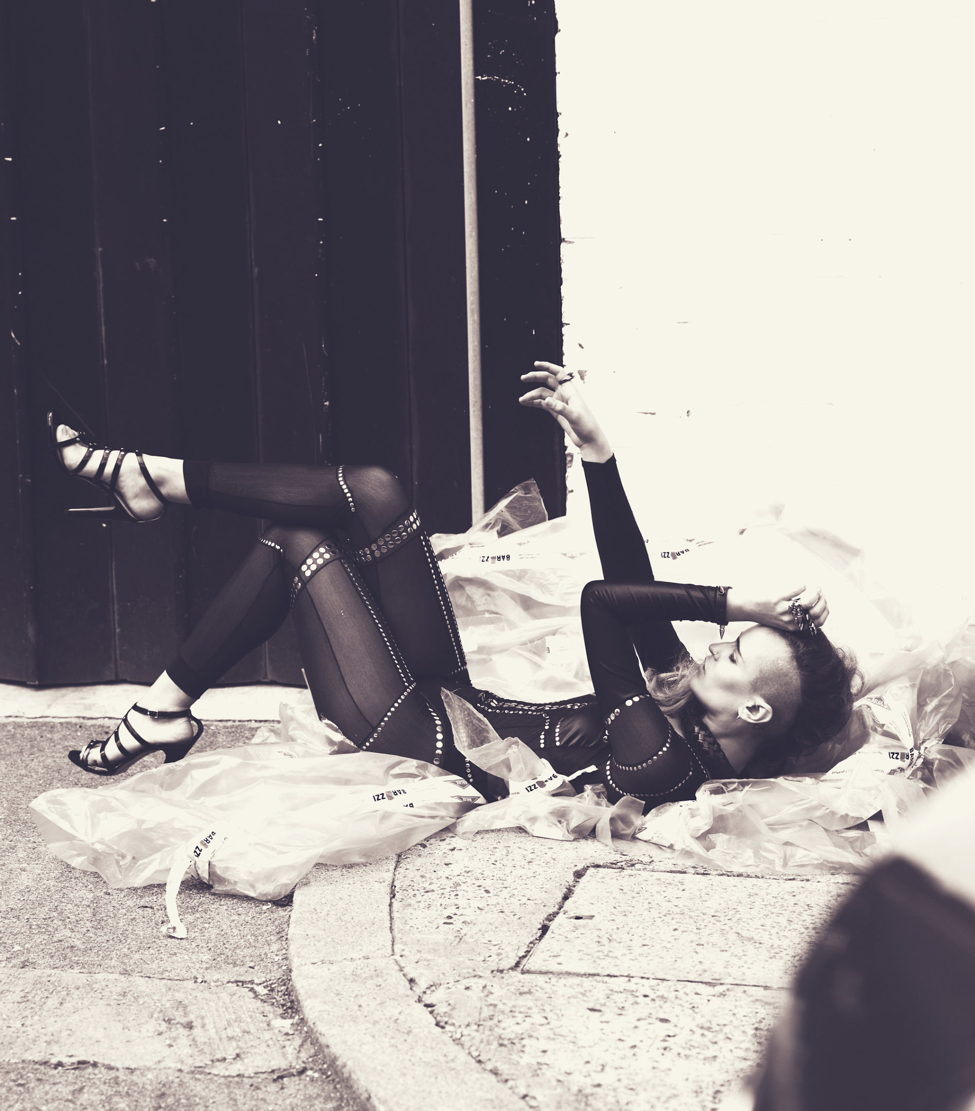 A woman in a black bodysuit and high heels lies on the pavement surrounded by translucent plastic bags in a monochrome outdoor shot.