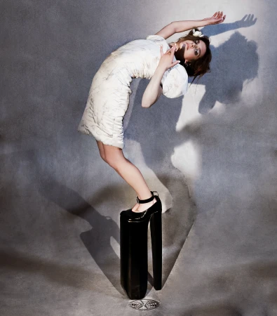 Woman in white dress arching backward while balancing on extremely high black platform shoes against a grey textured studio backdrop.