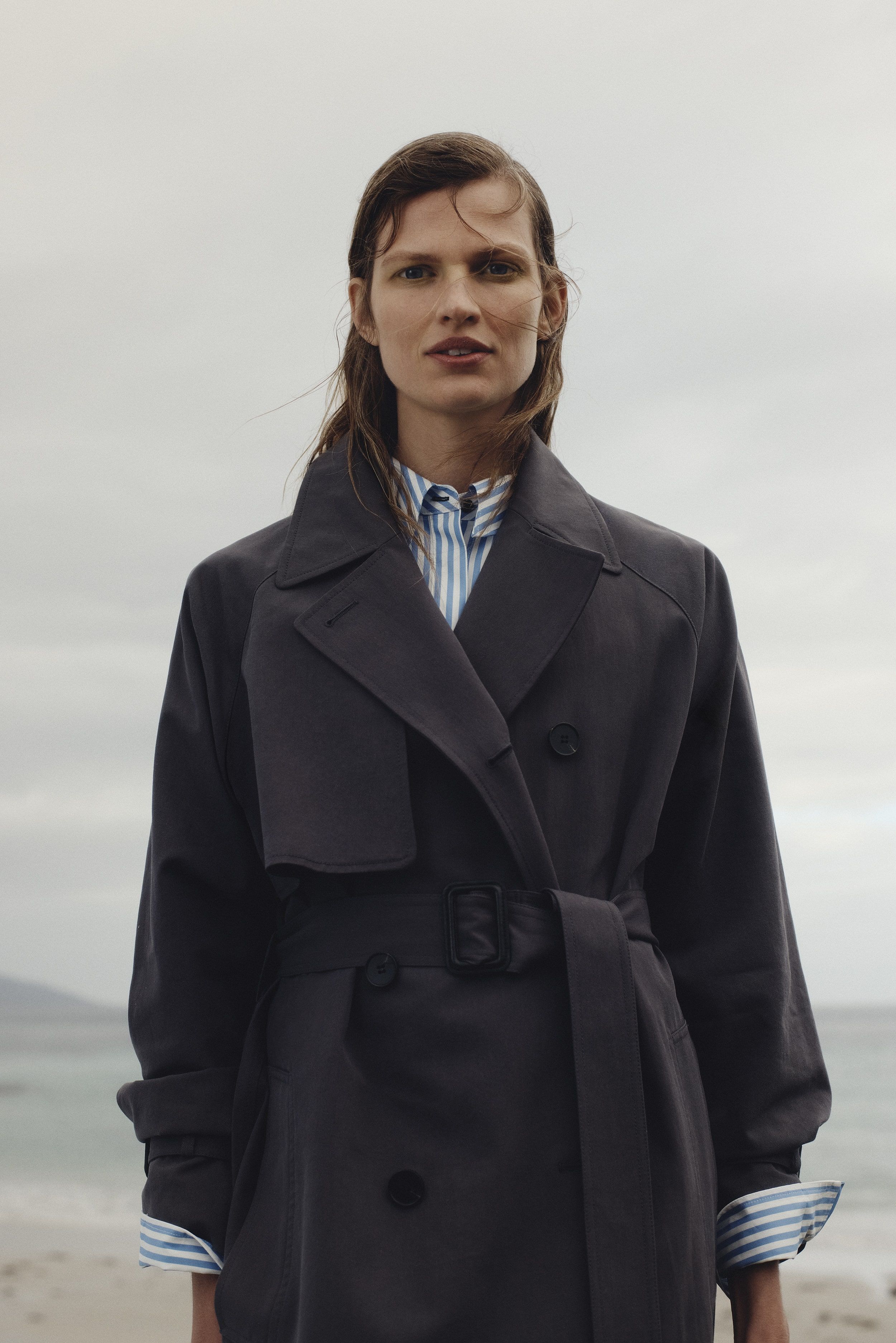 A woman in a belted trench coat poses on a beach with a blue and white striped shirt beneath, under a cloudy sky.
