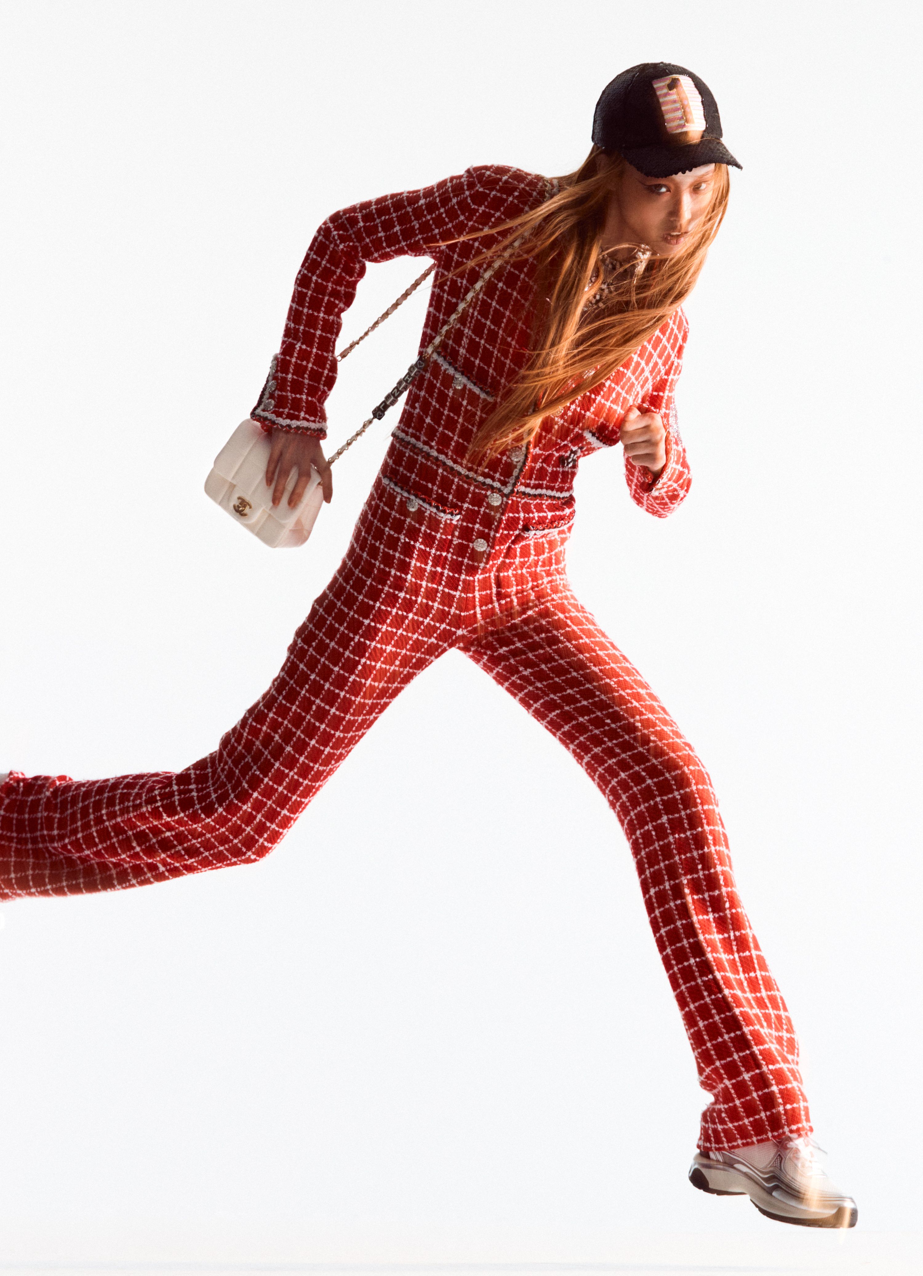 A female fashion model leaps against a white backdrop, showcasing a red and white checkered tweed suit and a small white purse.
