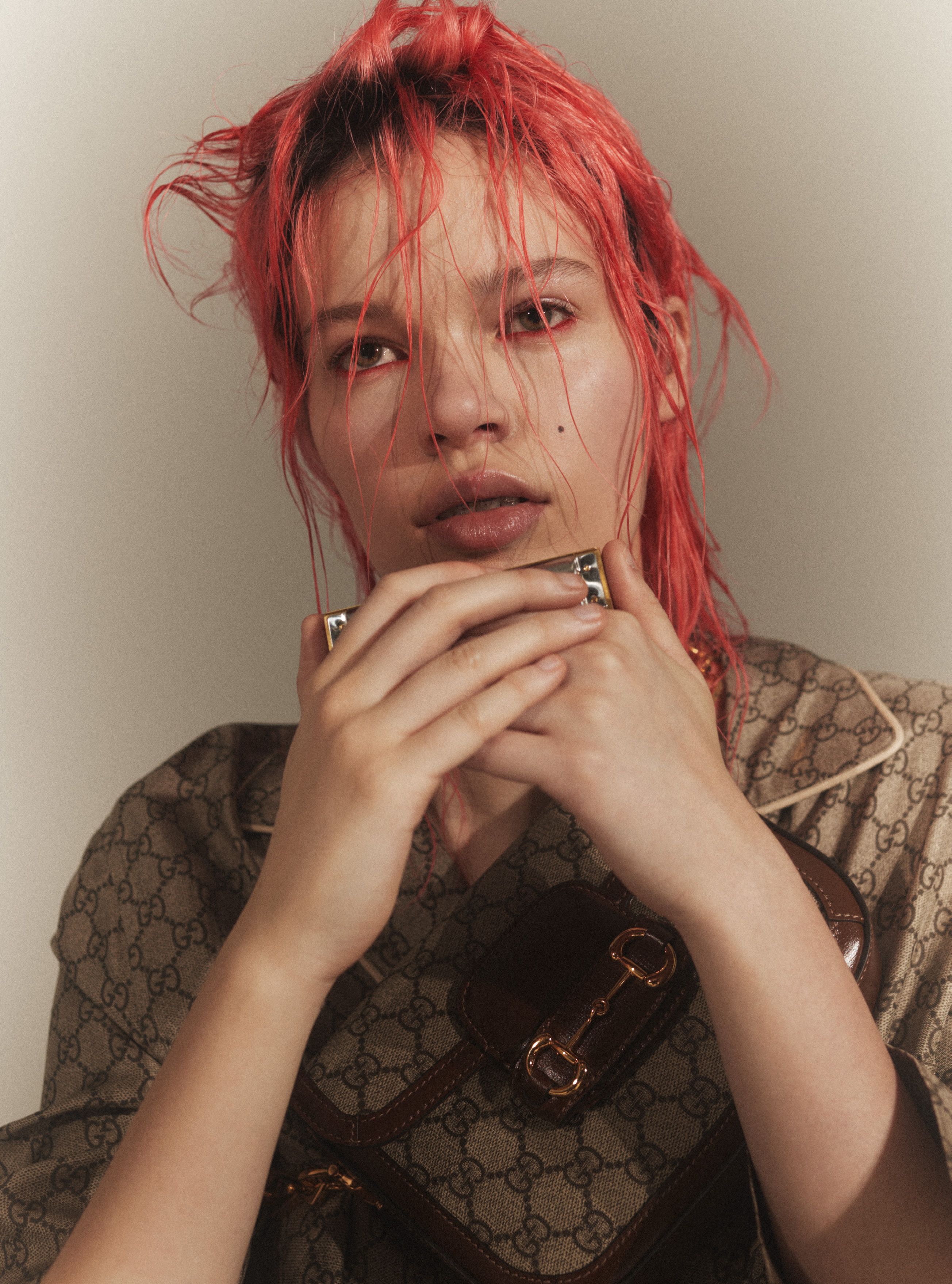 Close-up of a person with pink hair wearing a patterned shirt and holding a metal object near their mouth against a neutral background.