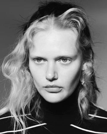 Close-up portrait of a model with blonde-streaked dark hair and a black turtleneck against a grey background