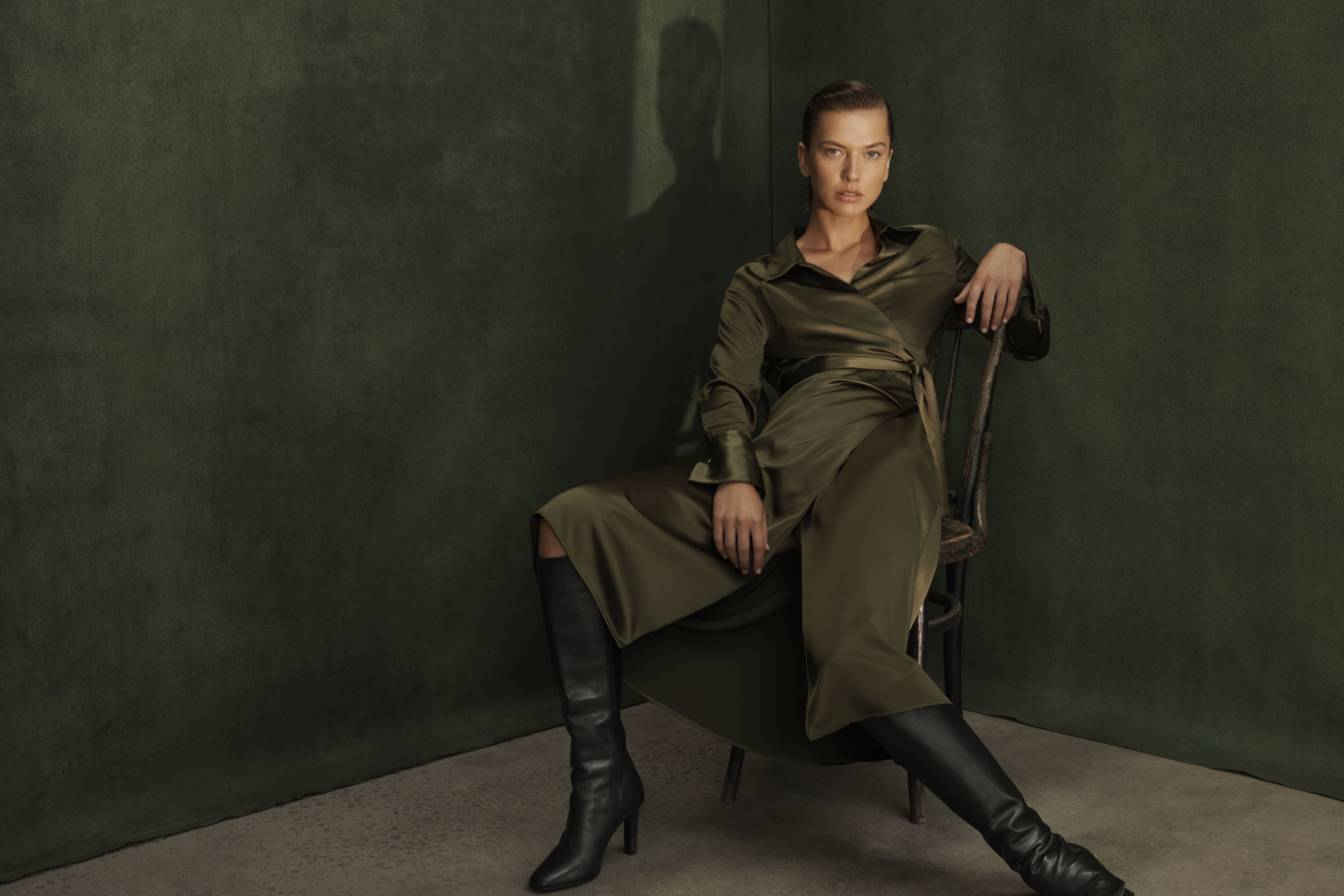 Woman seated in olive dress and boots in studio with soft lighting and shadows. Fashion photography promoting textile and style.