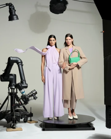 Two women model coordinated lilac and beige outfits in a studio, with a green handbag, under studio lighting and captured by a robotic camera.