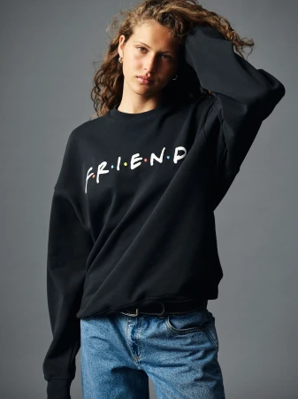 A woman with curly hair wearing a black sweatshirt and blue jeans against a grey studio backdrop in Sydney.