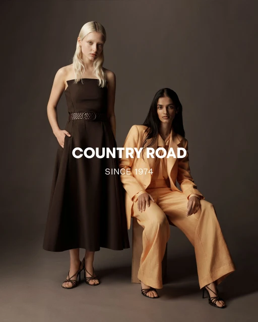 Two fashion models pose in Country Road apparel: one in a brown dress, the other in a peach suit, against a neutral studio backdrop.
