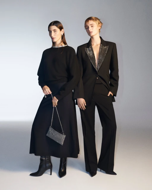 Two women pose in black outfits against a gradient background. The woman on the left carries a glittery clutch. Fashion shoot.