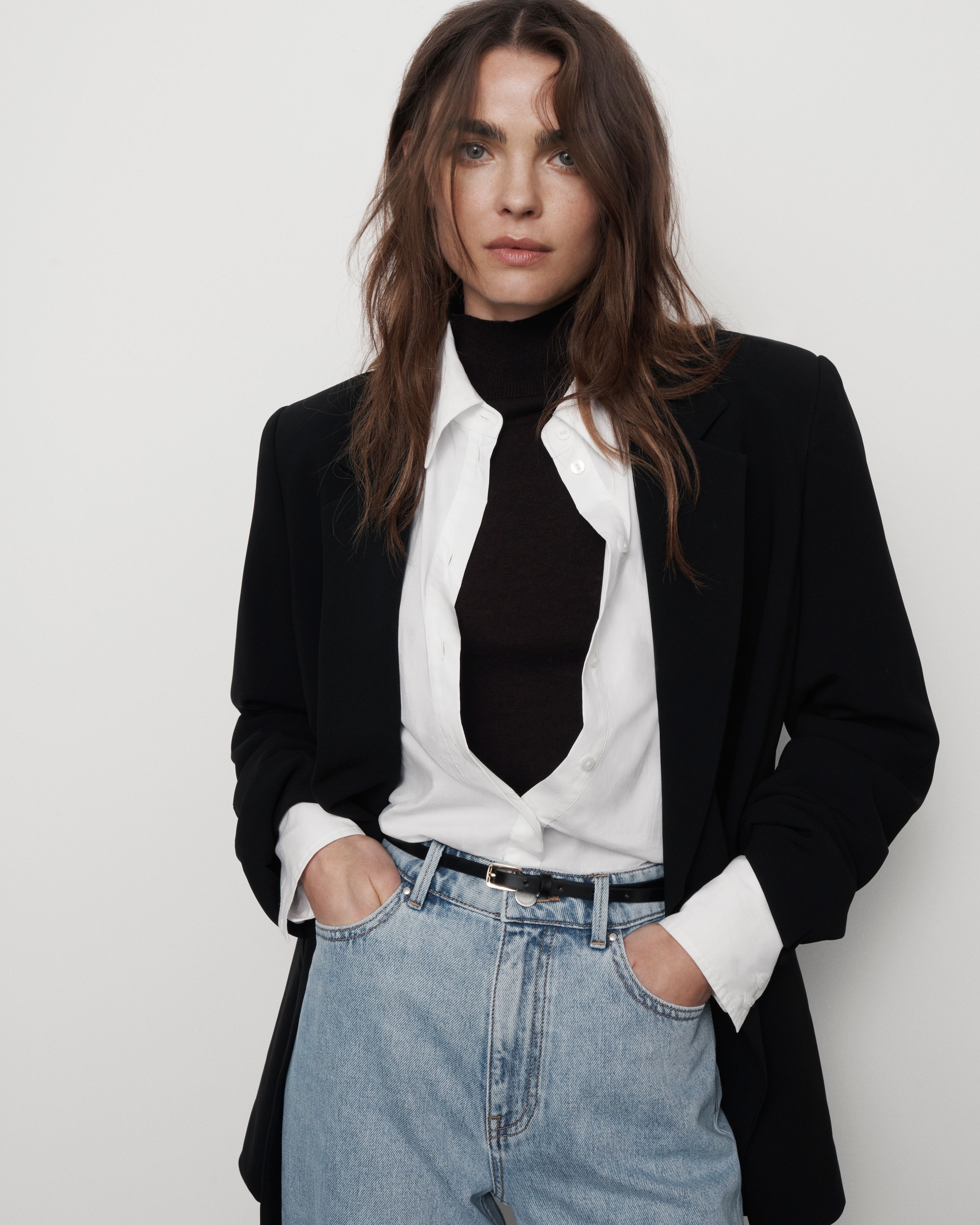 Woman wearing a black blazer over a white shirt and black turtleneck with blue jeans against a neutral background