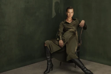 Model seated on a wooden chair wearing an olive silk wrap dress and black leather boots against a dark textured green wall