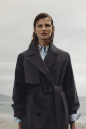 Woman in dark grey belted trench coat over blue and white striped shirt standing near an overcast coast