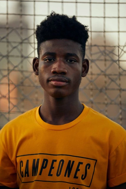 Close-up of a young Black man with an afro, wearing a yellow t-shirt with "CAMPEONES 1992" printed on it, standing against a blurred background.