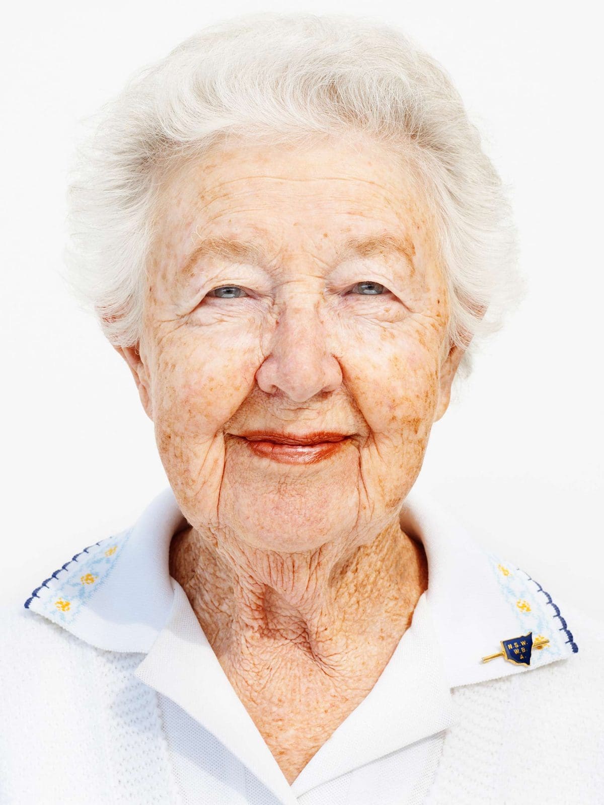 Portrait of a smiling elderly woman with white hair against a bright white background, wearing a white collared shirt and a pin.