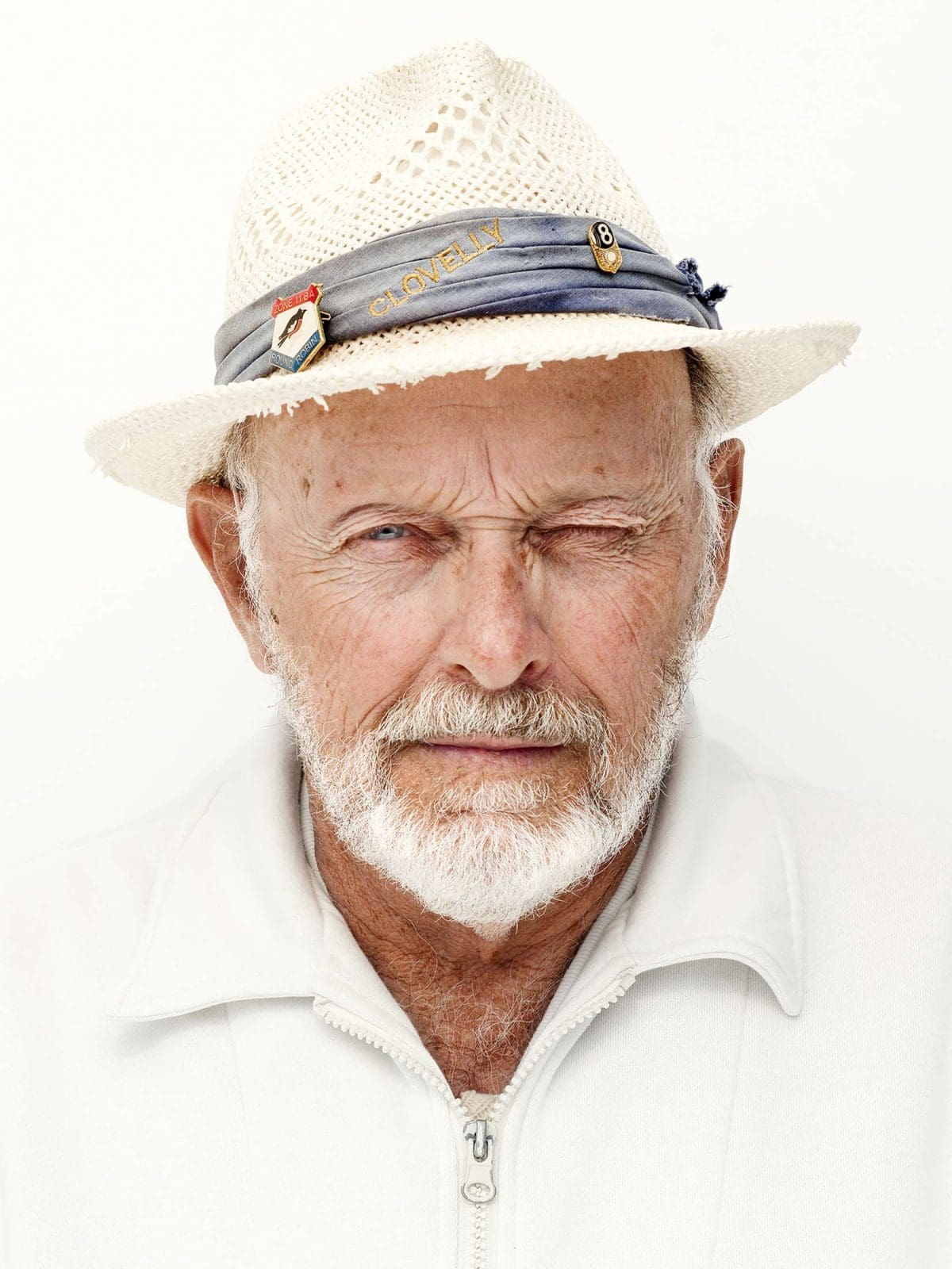Close-up portrait of an elderly man with a white beard wearing a woven hat with a blue band, winking against a white background.