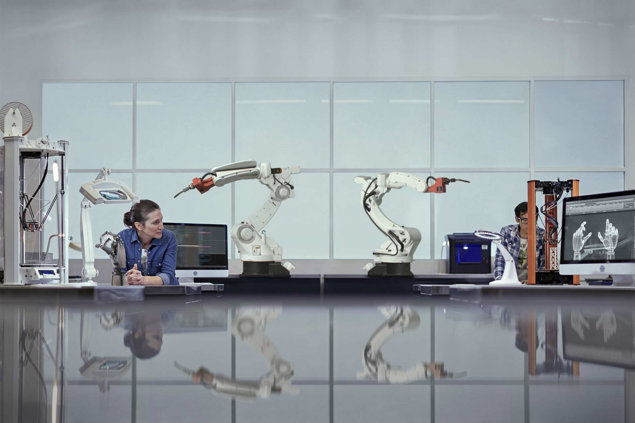 Two engineers work with robotic arms and 3D printers in a bright, modern laboratory with a large table and reflective floor.