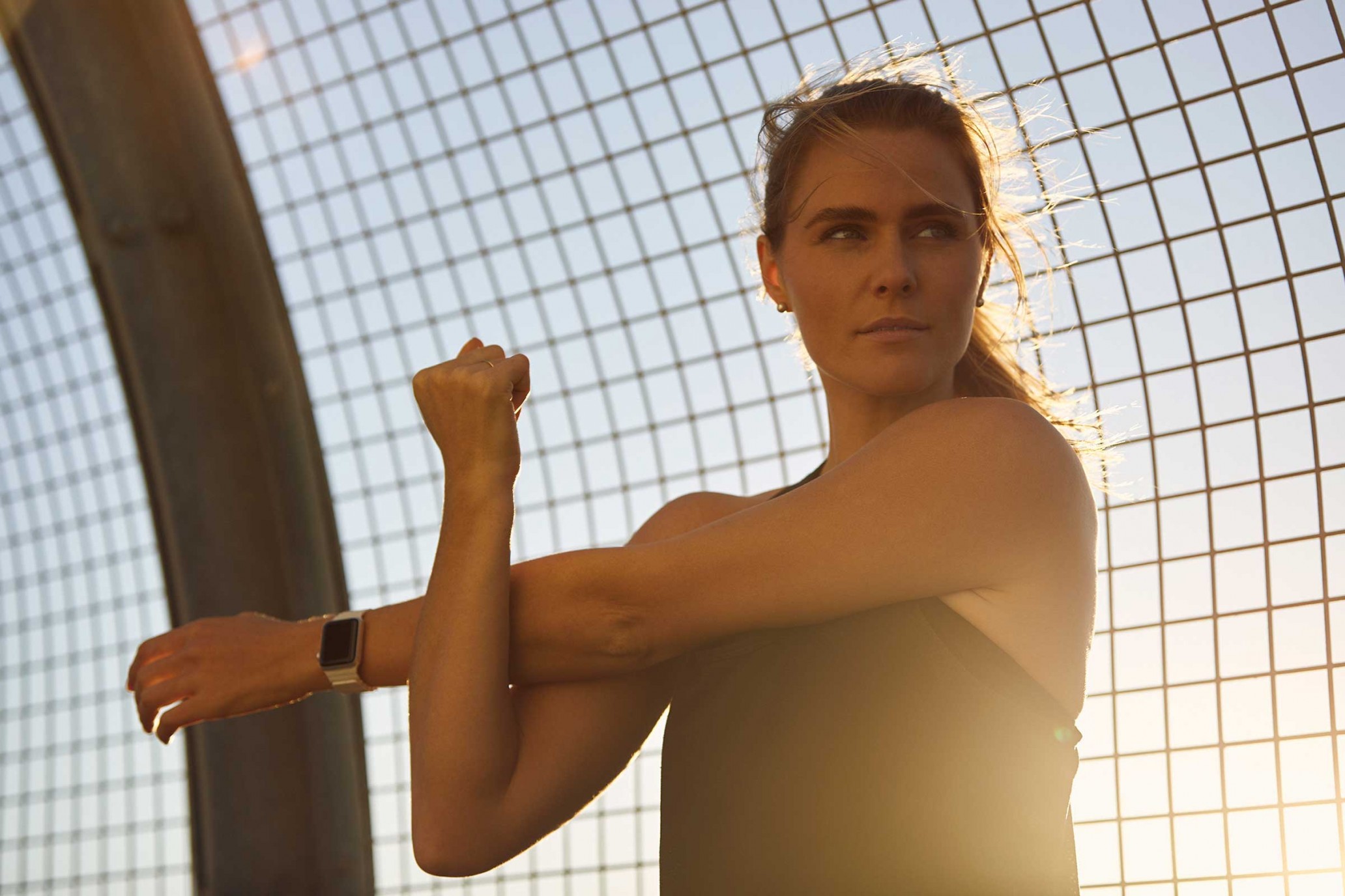 Woman stretching arms outdoors against a chain-link fence in sunlight, wearing a black tank top and a smartwatch.