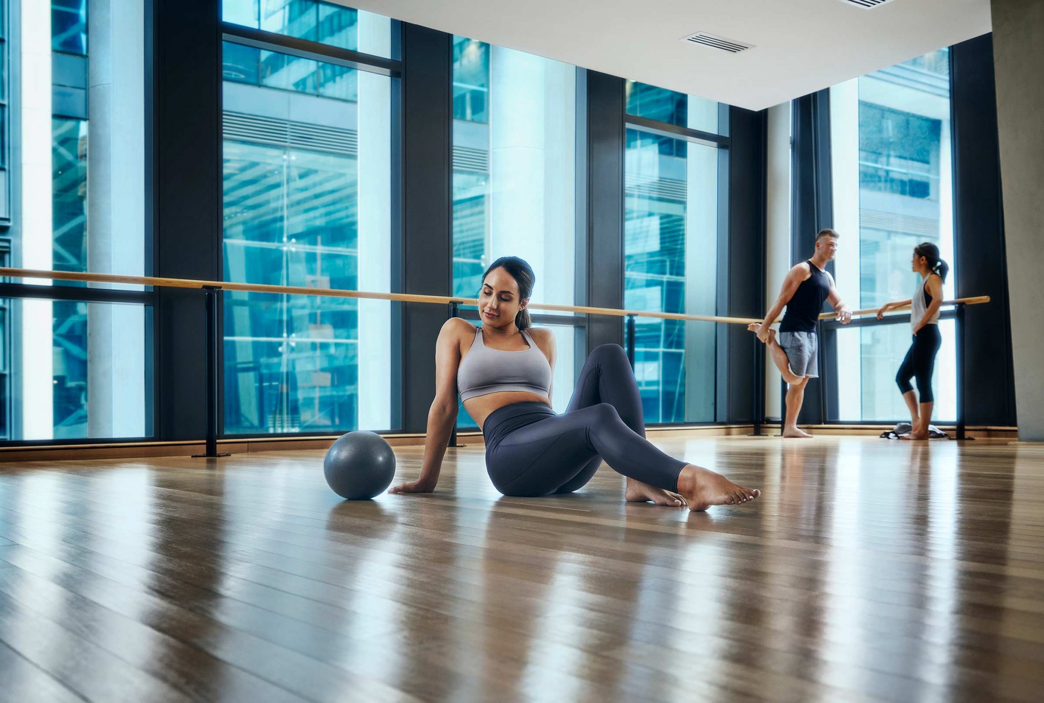 A woman in workout clothes sits on a wood floor next to a pilates ball in a bright, modern fitness studio with large windows.