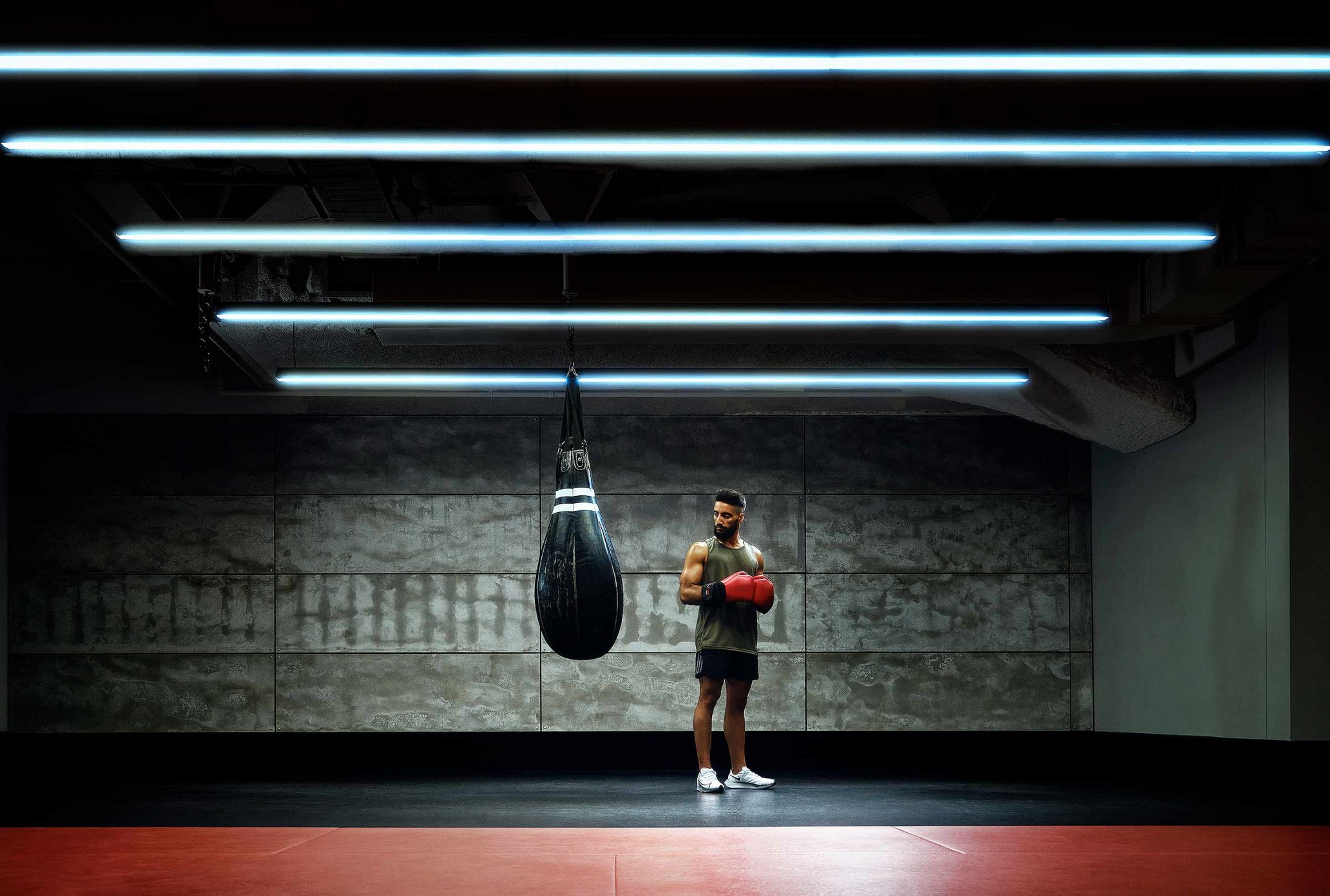Man in sportswear stands near a black punching bag in a gym with bright lighting, red boxing gloves, and white sneakers, looking fit and determined.