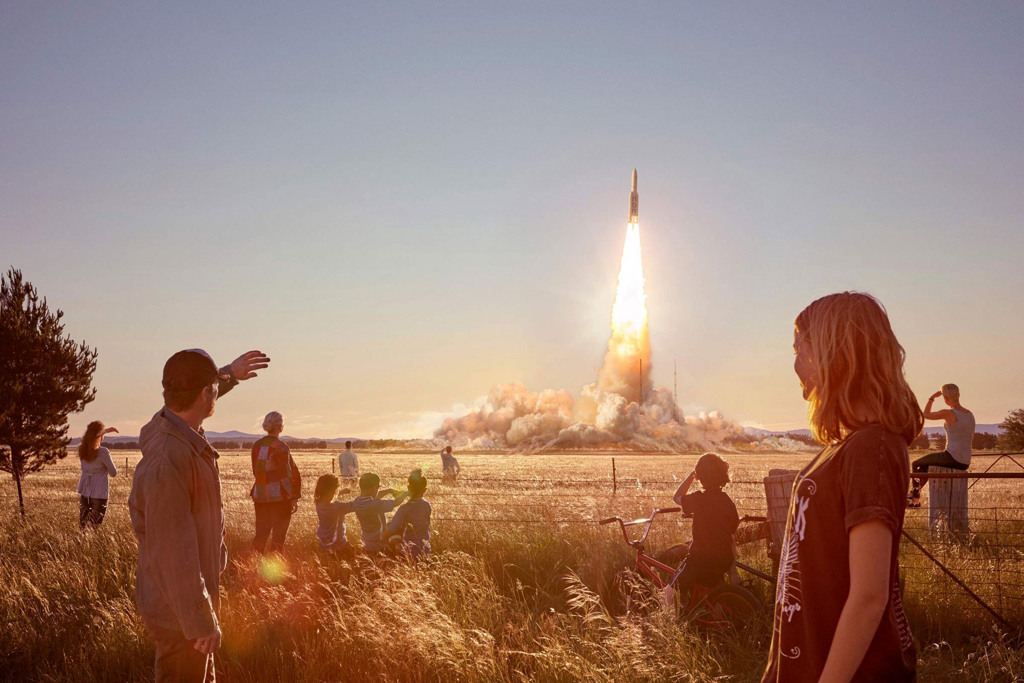 A rocket launches in a rural field observed by a diverse group of adults and children silhouetted against the sunset sky and golden grasses.
