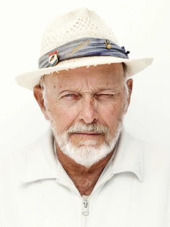 Close-up portrait of an elderly man with a white beard wearing a straw hat and white zip jacket against a plain white background.
