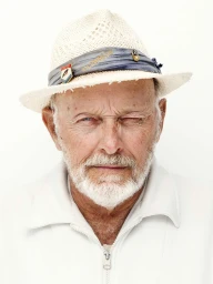 Close-up portrait of an elderly man with a white beard wearing a straw hat and white zip jacket against a plain white background.