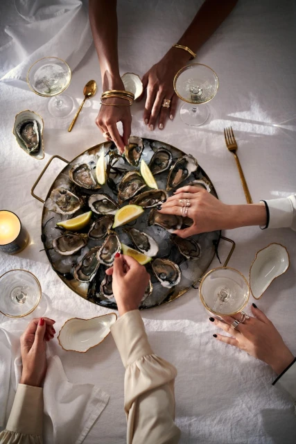 Overhead view of several women enjoying oysters and champagne on a white tablecloth-covered table in bright, diffused lighting.